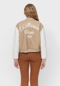 Chaqueta bomber beige con mangas blancas, con texto bordado "Le Marais Paris 1981" en la espalda. Pantalones marrones con estampado visibles debajo.
