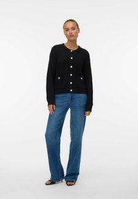 Black knitted cardigan with round neckline, button closure, and two front pockets. Paired with light blue wide-leg jeans and black sandals.