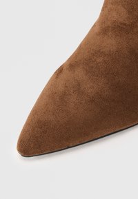 Punta afilada de una bota de ante marrón que muestra una cuidada textura suave sobre un fondo blanco.