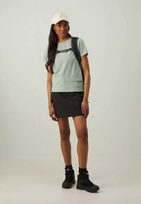 Ανοιχτό πράσινο μπλουζάκι με τσέπη στο μπροστινό μέρος, συνδυασμένο με μαύρη φούστα. Το μοντέλο φοράει μπεζ καπέλο και μαύρο σακίδιο πλάτης, με μαύρα παπούτσια.