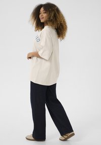 T-shirt beige surdimensionné à manches courtes, avec des lettres bleues, associé à un pantalon en denim sombre ample et des baskets de couleur claire.