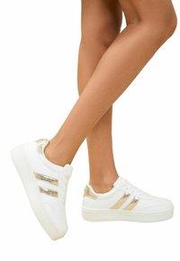 White sneakers with gold accents and textured details, featuring three stripes, a perforated toe box, and a thick white sole.