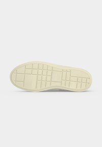Semelle d'une chaussure décontractée beige avec une semelle à motif quadrillé et une légère texture, présentée sur un fond gris clair uni.