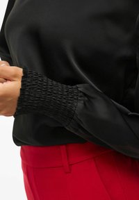 Blusa nera in tessuto morbido; presenta polsini arricciati e un leggero lucido; abbinata a pantaloni tailleur rossi per un contrasto.