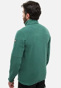 Veste en polaire verte avec un col haut, dotée d'un patch logo sur l'épaule et de coutures contrastantes. Tissu doux au toucher.