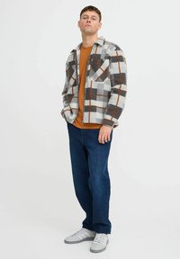Chaqueta de forro polar a cuadros gris y blanco con detalles en naranja, llevada sobre una camisa marrón, combinada con jeans azul oscuro y zapatillas grises.
