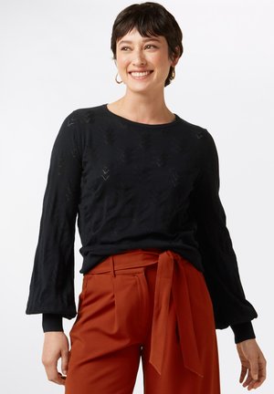 Maglietta nera a maniche lunghe con motivo a foglie testurizzato e polsini a costine, abbinata a pantaloni rustici a vita alta con cintura annodata.