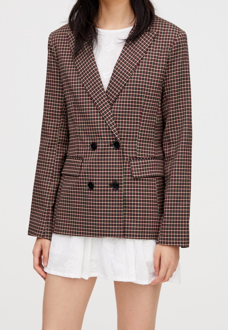 Femme portant un blazer croisé à carreaux marron sur une robe blanche en dentelle, montrée des épaules jusqu'à mi-cuisse.
