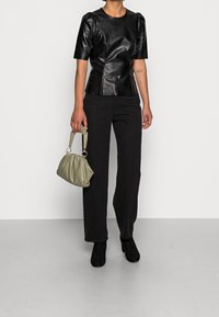 Person som bär en svart texturerad kortärmad topp, svarta byxor, svarta ankelskängor och håller en liten olivgrön rynkad handväska.