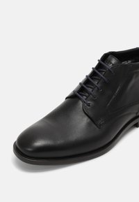 Chaussure habillée en cuir noir avec laçage fermé, bout affiné et talon bas, présentée sur un fond blanc uni.