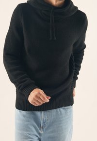 Svart stickad huvtröja med krage, ribbade manschetter och fåll. Texturen är tjock och mjuk, parat med ljusblå denimjeans.