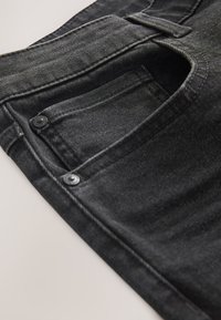 Nahaufnahme einer schwarzen Jeanshose-Tasche mit abgesteppten Details und Metallnieten auf hellem Hintergrund.