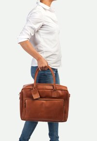 Bruine leren laptoptas met twee handvaten aan de bovenkant, een voorste ritsvak en een bagagelabel. Kenmerkt een gladde, gestructureerde afwerking en rechthoekige vorm.