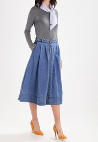 Jupe trapèze en denim avec fermeture boutonnée à l'avant, longueur mi-haute, couleur bleu clair, avec poches latérales, portée avec un haut gris à manches longues et des talons jaunes.