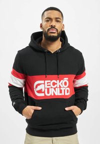 Crna i crvena dukserica s bijelim grafičkim logotipom. Ima prednji "kangaroo" džep, kapuljaču s čipkom i kontrastne crvene i bijele pruge na rukavima.