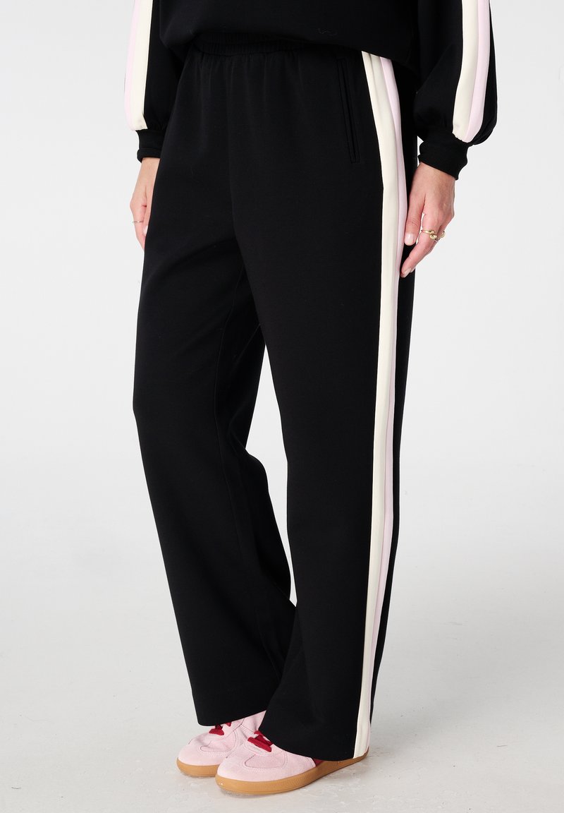 Pantalon de survêtement noir avec des rayures verticales roses et blanches, une taille élastique confortable et une coupe ample, assorti à des baskets roses.