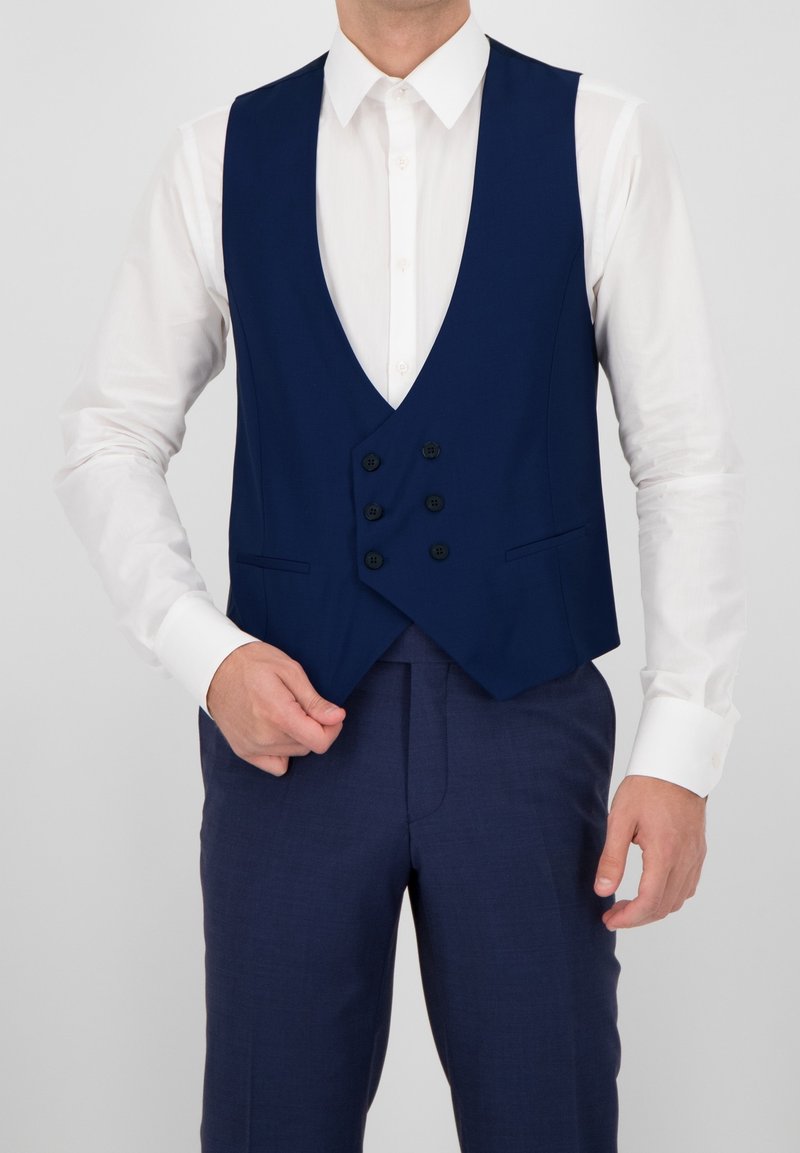 Hombre vistiendo un chaleco abotonado azul oscuro sobre una camisa blanca de vestir y pantalones a juego azul oscuro, ajustando el chaleco con una mano.