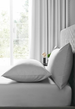 Zwei weiße Kissen auf einem grau gepolsterten Bett mit weißen Vorhängen und einer Glastür, die natürliches Licht hereinlässt.