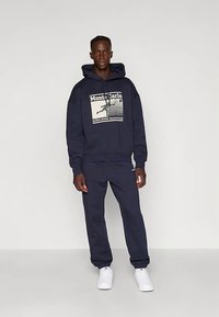 Φούτερ με κουκούλα σε ναυτικό μπλε και αντίστοιχο παντελόνι φόρμας, με εκτυπωμένο γραφικό «Monte Carlo» και «BJORN BORG Tennis Club» σε μπεζ χρώμα.