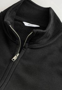 Schwarze Zip-Jacke mit silbernem Reißverschluss, hohem Kragen und sichtbarem inneren Fleece-Futter, mit Markenlabel "NEXT" im Kragen.