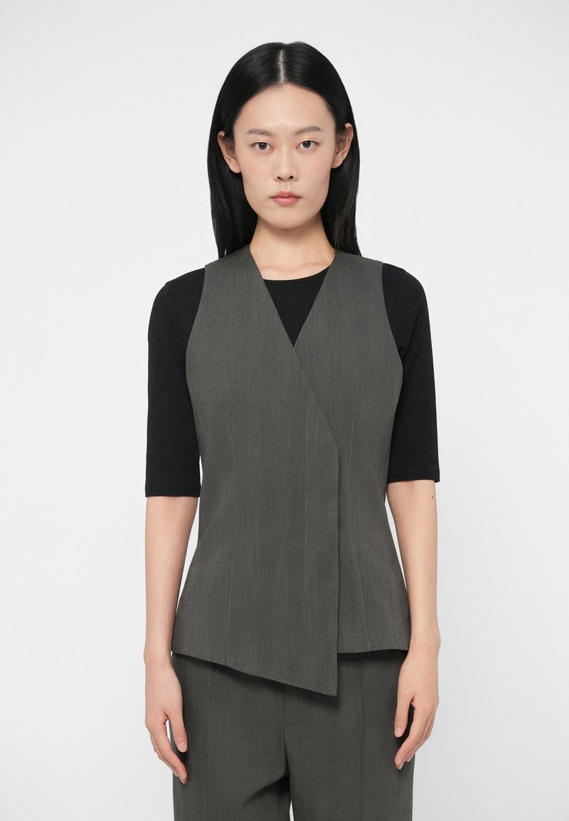 Sivý V-čkový bezrukávový sveter s prekrývajúcim sa dizajnom a vertikálnymi pruhmi, v kombinácii s čiernou priliehavou košeľou s hladkou text�úrou.