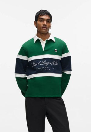Jeune homme portant un maillot de rugby vert, blanc et bleu marine avec texte "Karl Lagerfeld" et pantalon noir, debout devant un fond blanc.