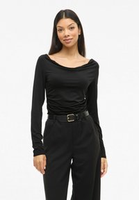 Zwarte langemouwen top met een gedrapeerde halslijn, gemaakt van een soepele stof. Gecombineerd met hoge zwarte broeken en een zwarte riem met een gouden gesp.