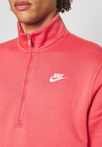 Röd polyestertröja med hög krage och halvdragkedja. Har en vit Nike-logotyp på vänster bröst och en slät yta.