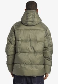 Man med olivgrön puffjacka med huva, visad bakifrån, ihop med mörkblå jeans.