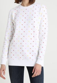 Felpa bianca a maniche lunghe con piccoli loghi "CK" multicolori su tutta la superficie, indossata da una persona con jeans scuri.