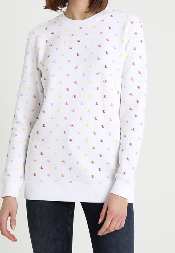 Felpa bianca a maniche lunghe con piccoli loghi "CK" multicolori su tutta la superficie, indossata da una persona con jeans scuri.
