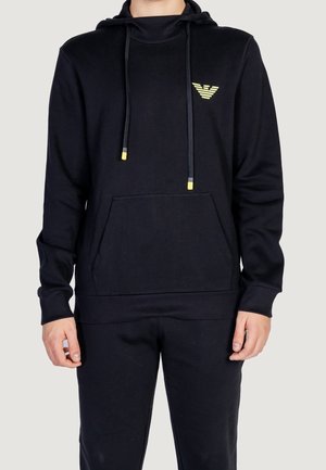 Man met een zwarte hoodie en broek met geel-accentueerde trekkoorden en een klein geel embleem op de borst, staande tegen een effen achtergrond.