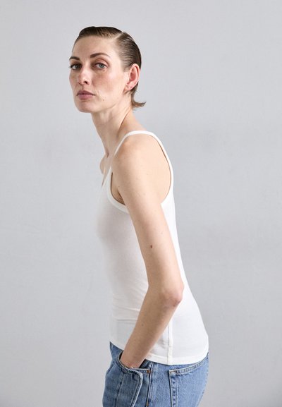 Bílý žebrovaný top na ramínka s úzkými popruhy, v kombinaci se světle modrými džínami s vysokým pasem. Modelka stojí bokem, ruce v kapsách.