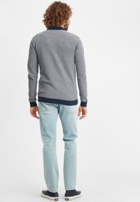 Pulover gri texturat cu guler bleumarin, cu manșete și tiv ondulate, asociat cu blugi de culoare albastru deschis. Aspectul general este casual și ajustat.