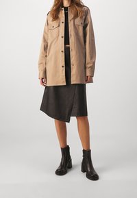 Tan button-up skjortejakke med to brystlommer, parret med en sort faux læder wrap nederdel og sorte ankelstøvler med kraftige hæle.
