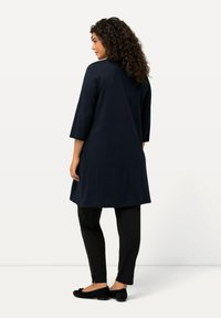 Marineblaues Tunika mit dreiviertellangen Ärmeln, der Stoff sieht glatt aus. Kombiniert mit schwarzen Hosen und schwarzen Ballerinas, die eine Schleifenakzent besitzen.