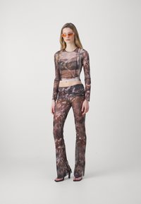 Transparante top met lange mouwen en flared broek in een bruine en gekleurde abstracte print, gecombineerd met oranje zonnebril en hoge sandalen.