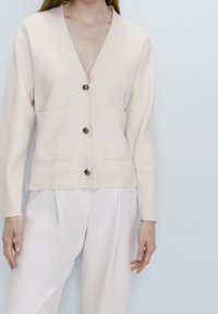Light beige cardigan with a v-neck, ribbed cuffs, and two front pockets. Features five brown buttons down the front. Texture appears soft.