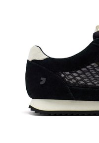 Zapatilla negra de ante con paneles de malla, detalles en gris claro y una suela de goma blanca. Presenta un logo negro texturizado en el lateral.