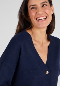 Cardigan blu navy con scollo a V profondo, caratterizzato da dettagli a coste e due piccole tasche, chiuso con un bottone color oro. Texture morbida.