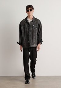 Veste en denim noir avec fermeture à boutons, deux poches poitrine et coupe décontractée. Portée sur une chemise blanche et assortie avec un pantalon et des chaussures noires.