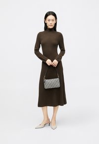 Καφέ ριχτό φόρεμα μακρύ μανίκι με ψηλό λαιμό. Το μοντέλο κρατά μια τσάντα με σχέδιο. Μπεζ γόβες slingback. Απλή, εφαρμοστή σιλουέτα.