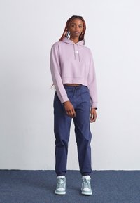 Haut de sweat à capuche cropped violet clair avec cordons, pantalon bleu marine fuselé et baskets teal avec accents blancs, dans un cadre minimaliste.