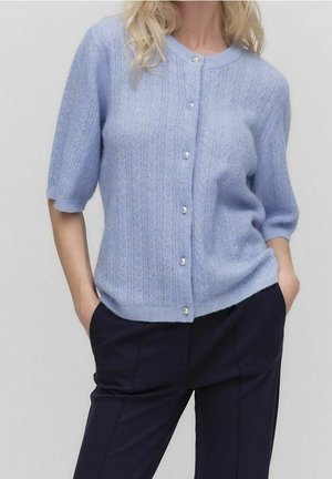 Kobieta nosząca jasnoniebieski, krótki sweter zapinany na perłowe guziki, w parze z dopasowanymi, ciemnogranatowymi spodniami, ręce w kieszeniach.