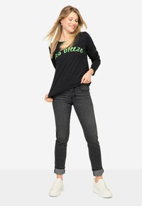 Camiseta negra de manga larga con texto en verde "brisa marina", combinada con pantalones vaqueros gris oscuro y zapatillas blancas. Diseño simple con un corte relajado.