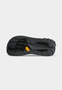 Semelle de chaussure noire robuste avec des crampons profonds et un petit logo hexagonal jaune sur un fond gris clair uni.