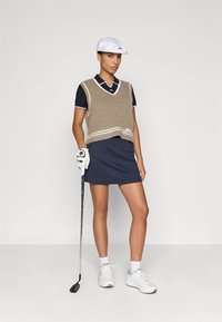 Bež pletena brezrokavnik z belimi detajli čez temnomodro polo majico, v kombinaciji s temnomodro rebrasto krilo. Bela kapa in golf rokavica, stoji z palico.