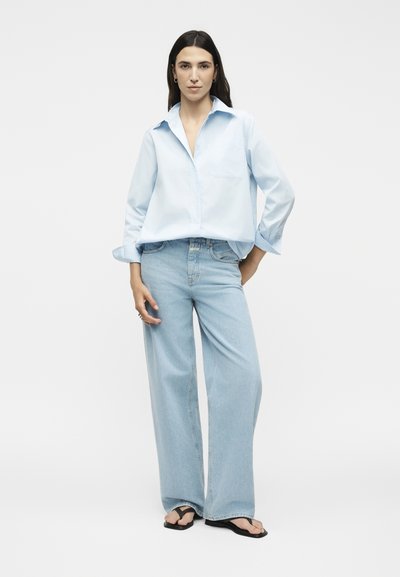Γυναίκα με μακριά σκούρα μαλλιά φοράει ένα ανοιχτό μπλε oversized πουκάμισο, ανοιχτό μπλε παντελόνι με φαρδιά μπατζάκια και μαύρα σανδάλια σε απλό φόντο.