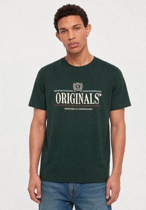 Jeune homme aux cheveux courts et bouclés portant un t-shirt vert foncé avec le texte "ORIGINALS Designed in Copenhagen" et un jean bleu sur un fond uni.