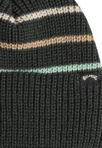 Bonnet en laine tricoté de couleur vert foncé avec des rayures vert clair, beige et blanche. Présente une texture côtelée et un petit patch logo noir.